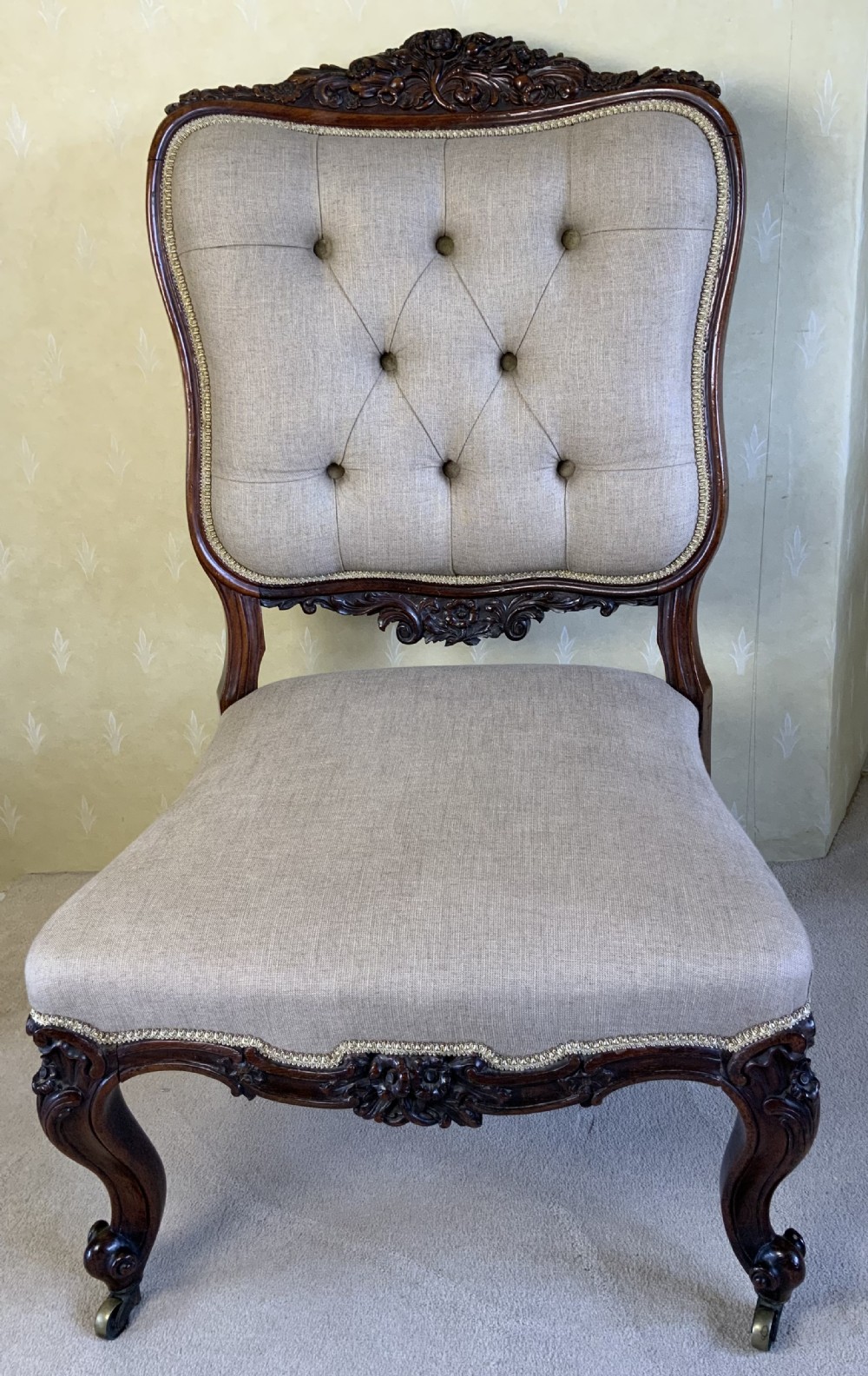 A mid-19th-century Victorian rosewood side chair featuring button-back beige linen upholstery, carved cresting rail, cabriole legs, and brass castors.