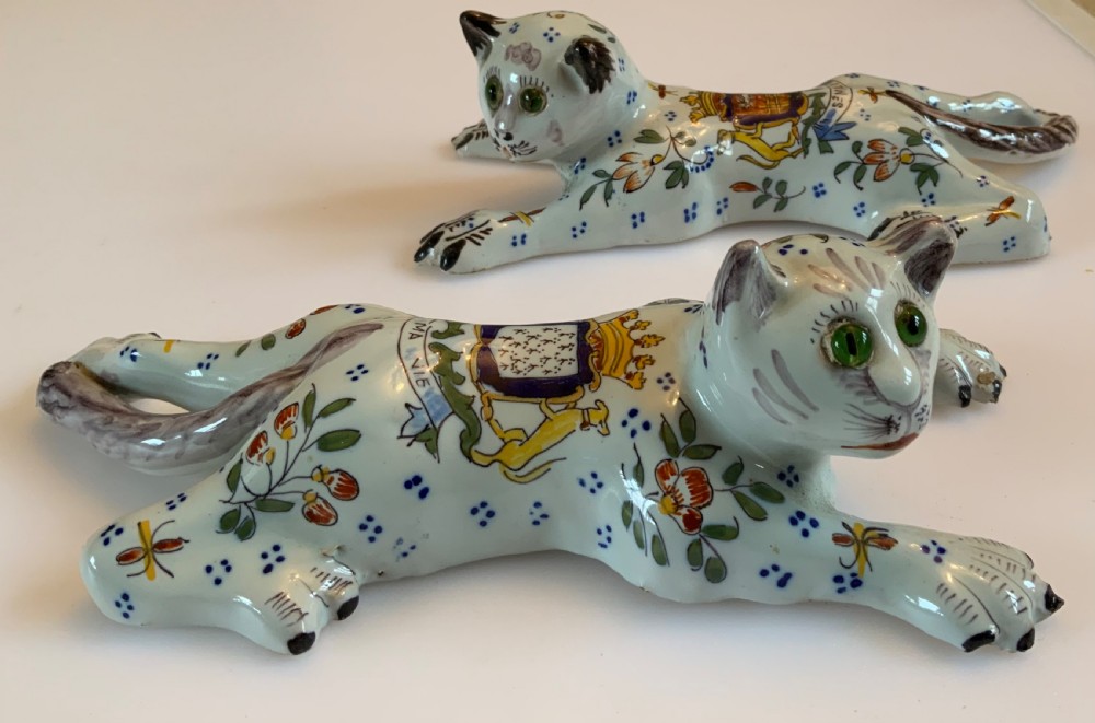 A pair of hand-painted tin-glazed ceramic cat wall pockets styled in recumbent poses with heraldic coat of arms motifs and green glass eyes.