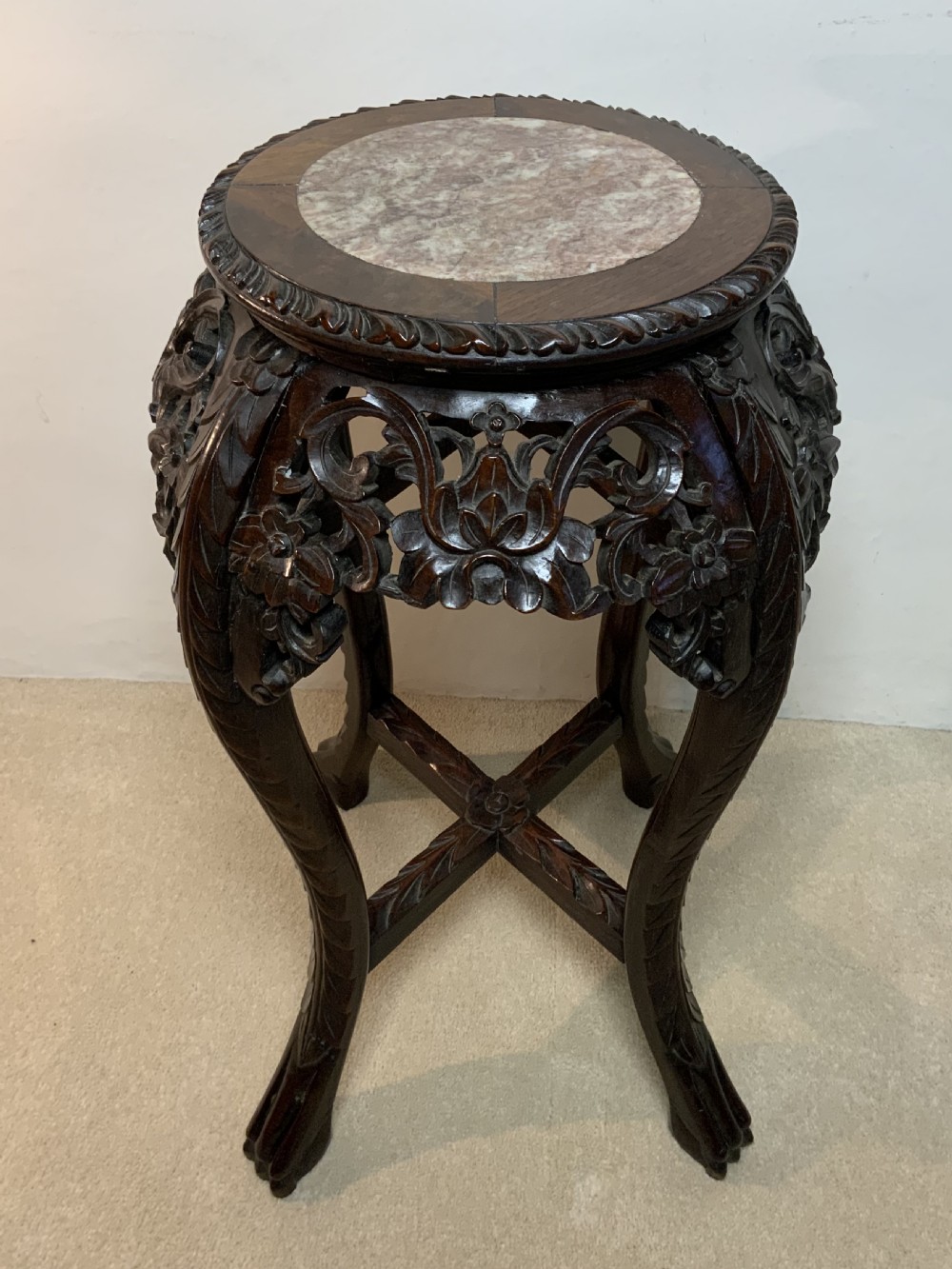A dark hand-carved hardwood round plant stand featuring an inset circular marble top, a pierced floral apron, cabriole legs, and an X-shaped cross stretcher.