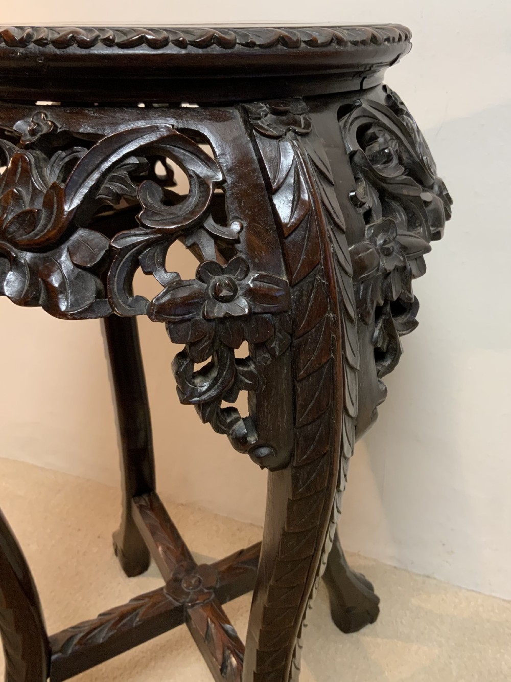 A dark hand-carved hardwood round plant stand featuring an inset circular marble top, a pierced floral apron, cabriole legs, and an X-shaped cross stretcher. View V