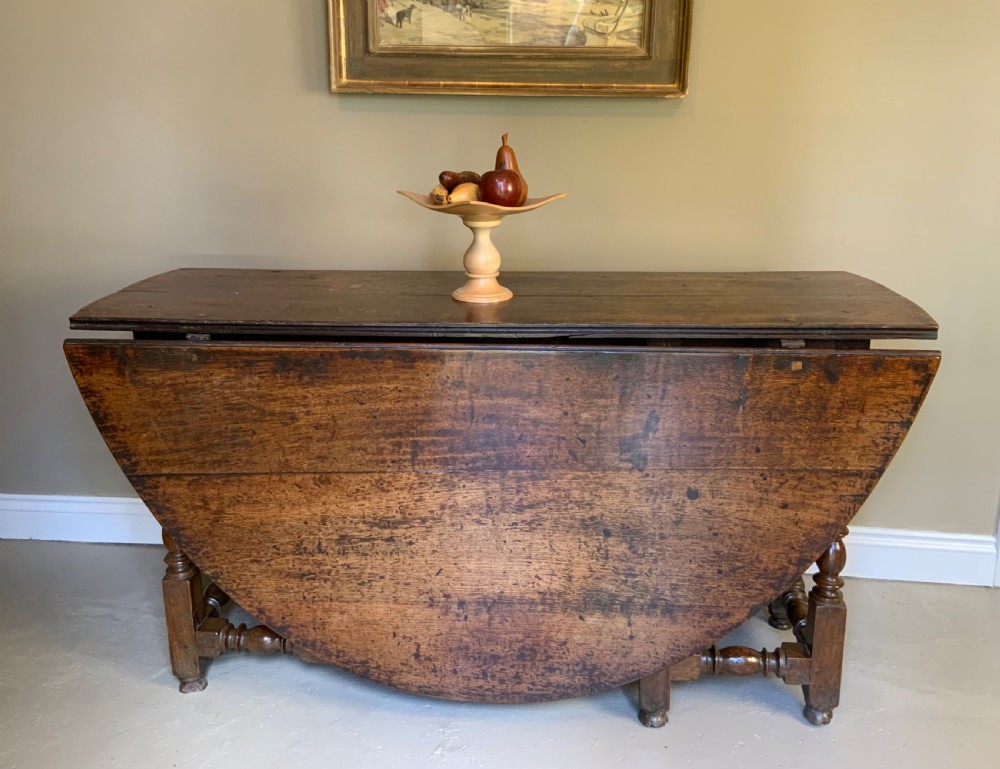 A large 17th-century solid oak gateleg dining table featuring an oval drop-leaf top and turned wooden legs. View VI