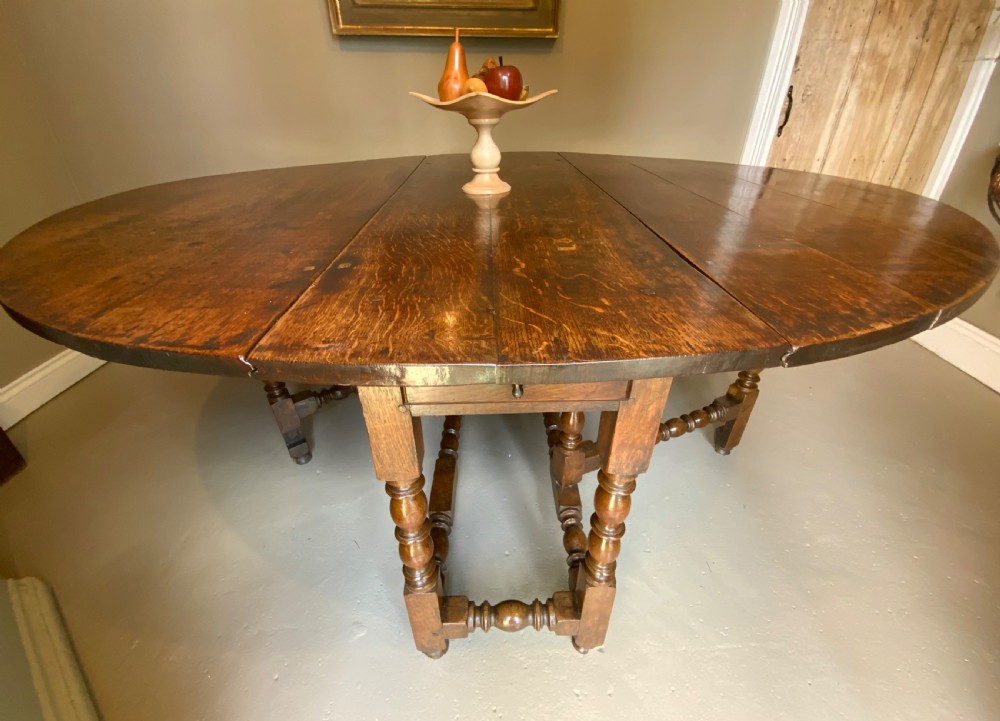 A large 17th-century solid oak gateleg dining table featuring an oval drop-leaf top and turned wooden legs.