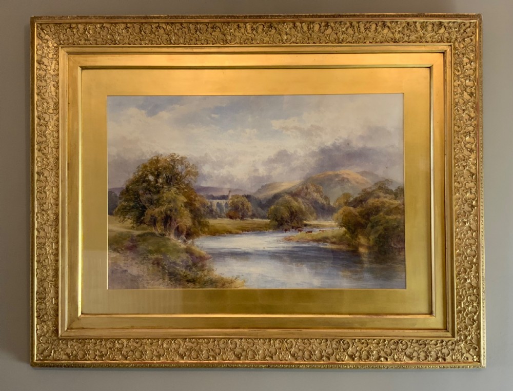 A framed 19th-century watercolour painting depicting a river running through a green landscape with trees and distant hills, set within an ornate gold gilt frame.