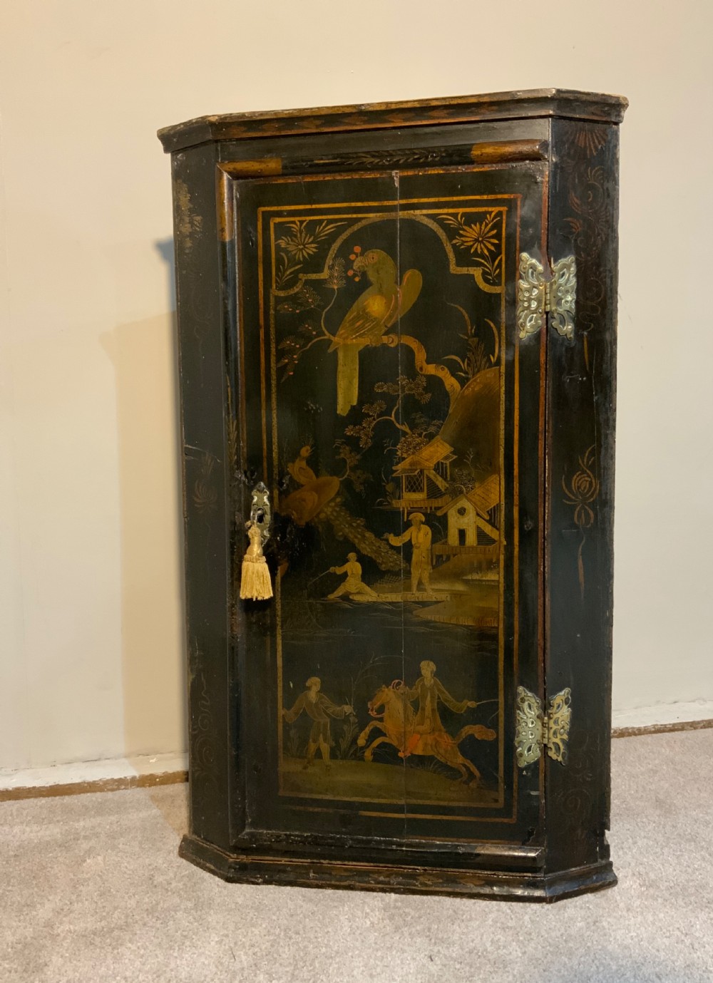 An 18th-century black lacquered corner cabinet featuring gilt chinoiserie artwork of figures, architecture, and a parrot, fitted with decorative brass hinges.