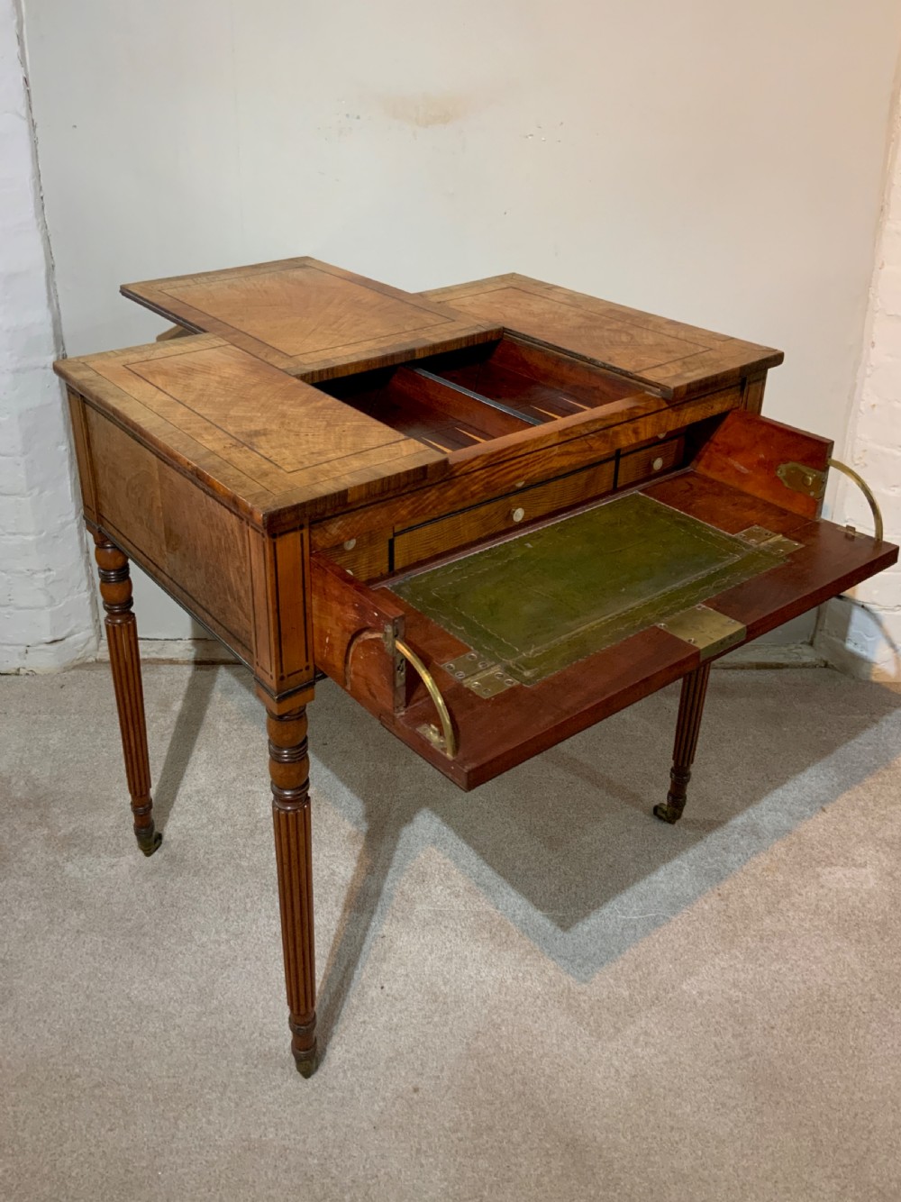 An antique mahogany Regency games and writing table featuring reeded legs, brass handles, a pull-out drawer with a green leather insert, and a reversible top revealing a game board compartment.
