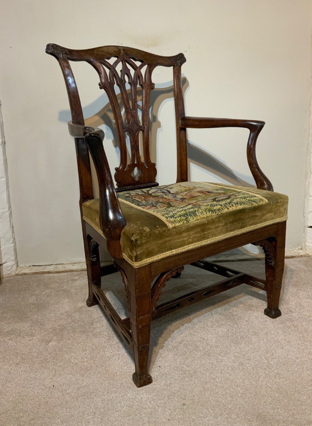 A Georgian dark mahogany armchair featuring an intricately carved pierced back splat, open curved arms, and a green velvet seat with an embroidered tapestry panel.