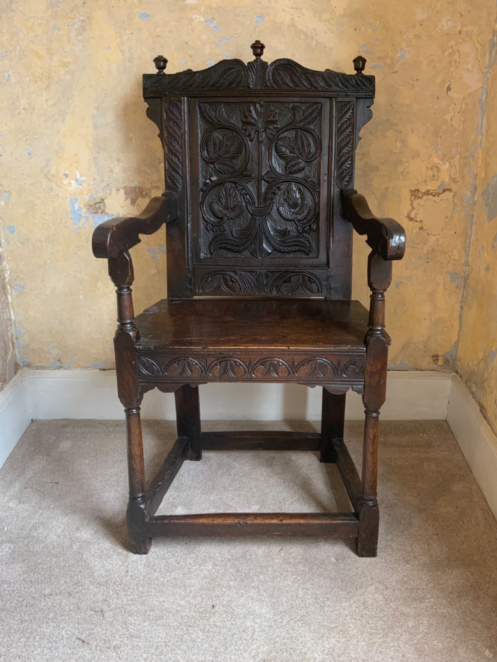 A dark oak 17th-century wainscot armchair featuring a high carved back panel with botanical motifs, turned front legs, and turned crest finials.