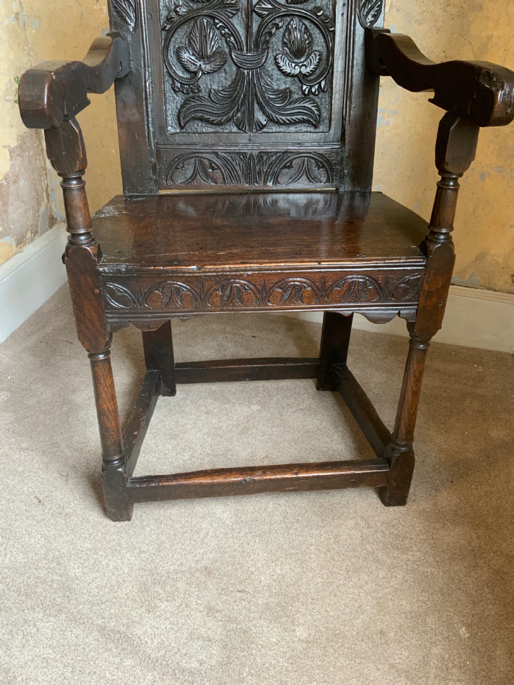 A dark oak 17th-century wainscot armchair featuring a high carved back panel with botanical motifs, turned front legs, and turned crest finials. View II