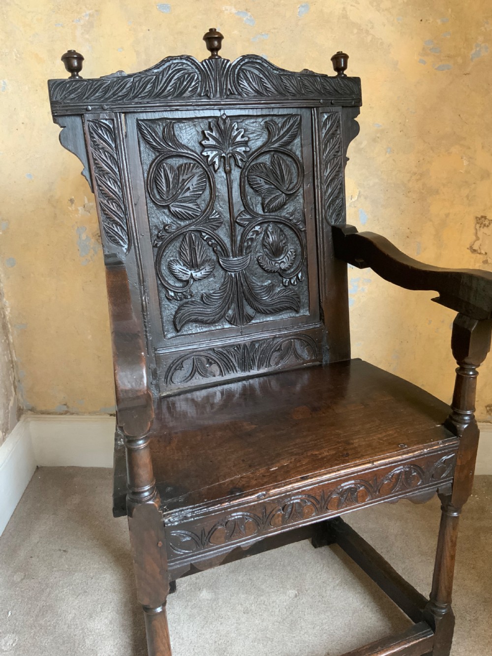 A dark oak 17th-century wainscot armchair featuring a high carved back panel with botanical motifs, turned front legs, and turned crest finials. View III