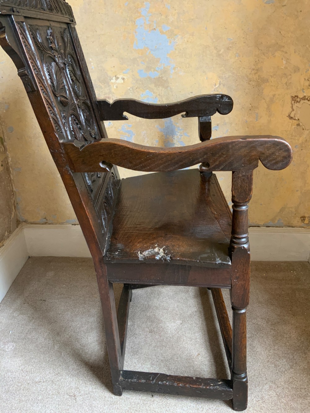 A dark oak 17th-century wainscot armchair featuring a high carved back panel with botanical motifs, turned front legs, and turned crest finials. View IV