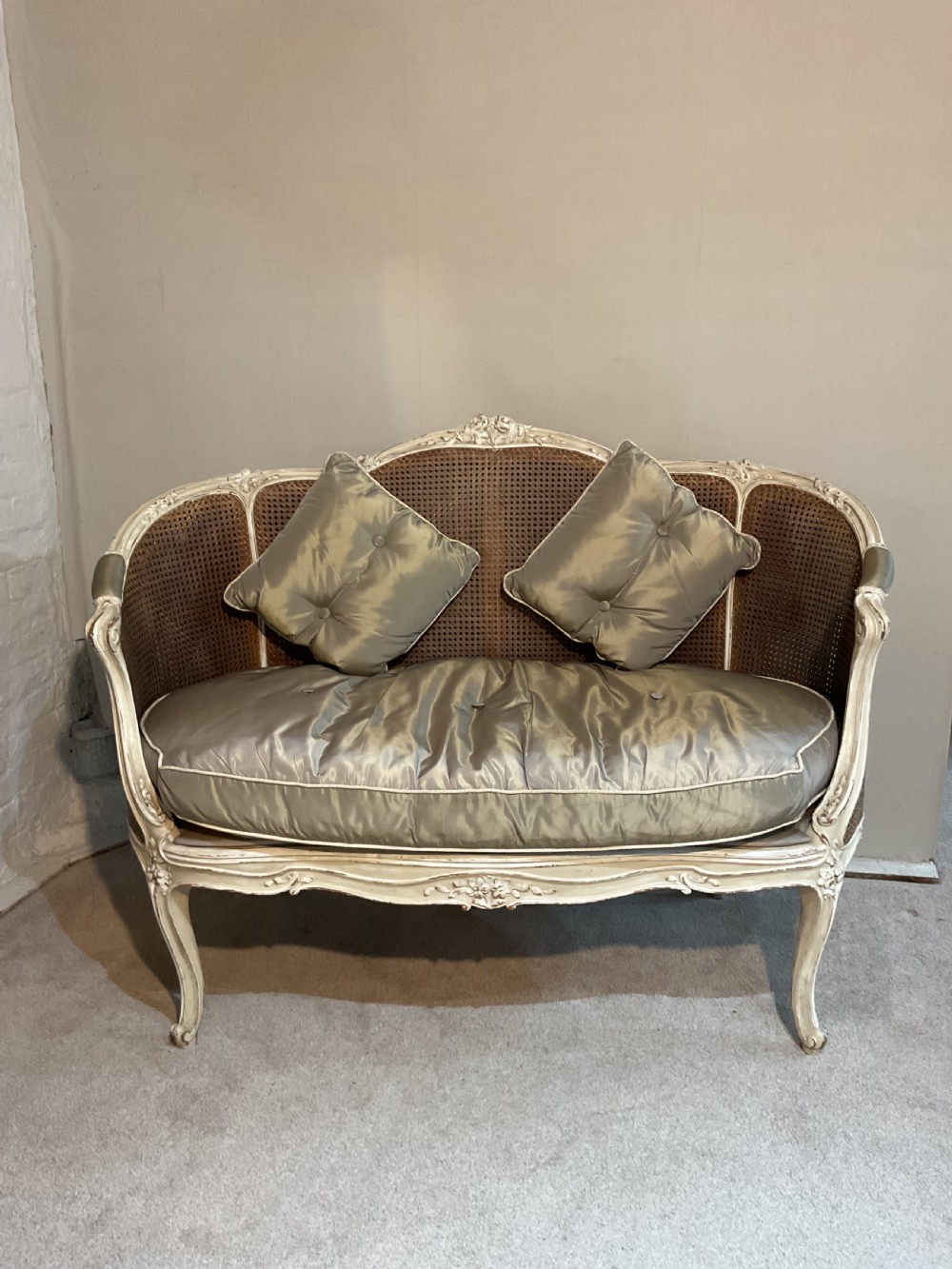 A 19th-century French bergere settee featuring a painted carved wooden frame with woven cane back panels and a champagne-coloured upholstered seat cushion with two accent pillows.