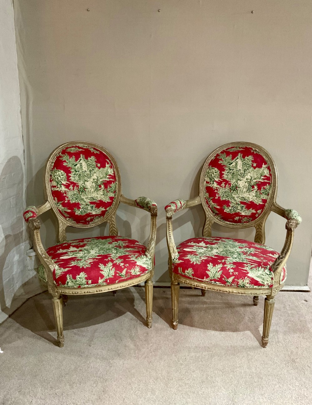 A pair of 18th-century French Louis XVI open armchairs featuring oval backrests, carved fluted legs, distressed gilding, and red patterned textile upholstery.
