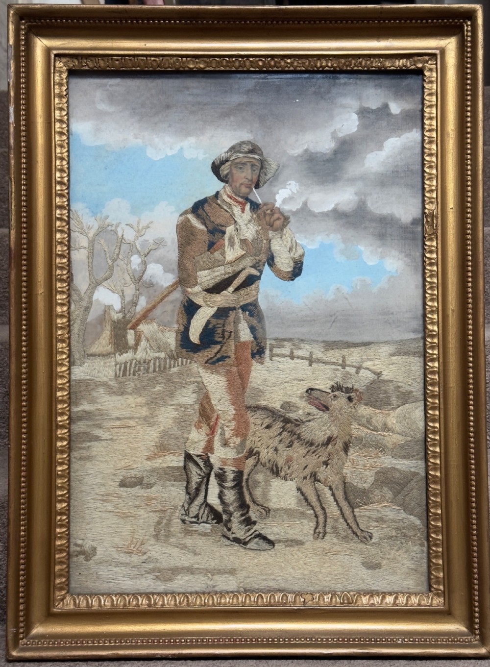 An antique late Regency long stitch silk embroidery depicting a country huntsman with his dog, framed in an ornate giltwood frame.
