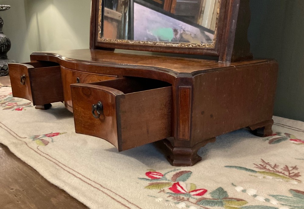 A George II period dark walnut toilet mirror featuring an arched adjustable swing frame set above a shaped three-drawer base with brass handles. View II