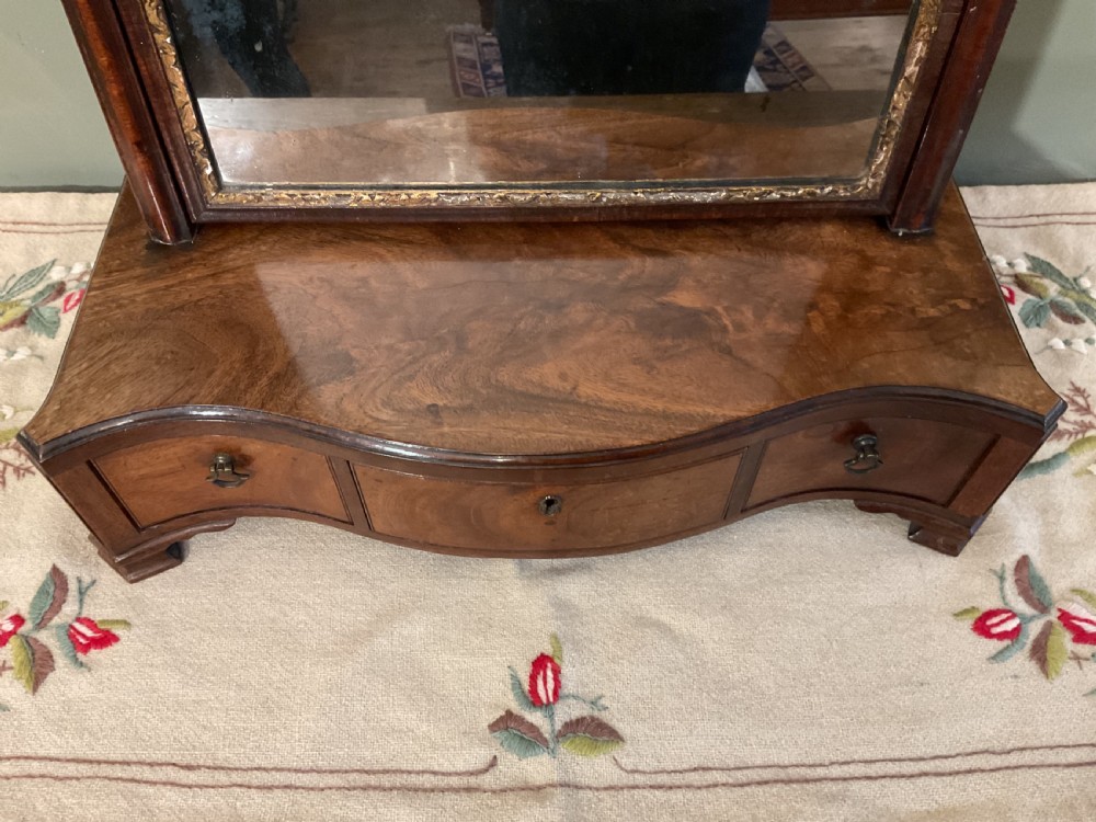 A George II period dark walnut toilet mirror featuring an arched adjustable swing frame set above a shaped three-drawer base with brass handles. View V