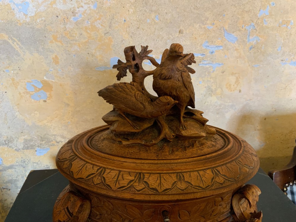 An antique carved wooden casket featuring a detailed top finial with two sculpted birds perched on foliage, sitting on carved feet with a keyhole in the front base. View V