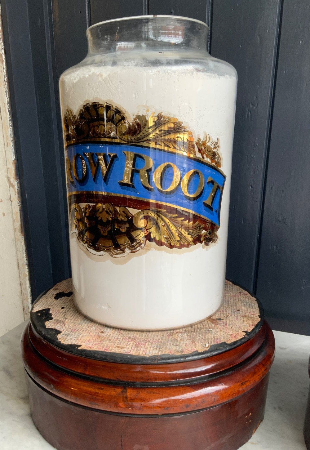 A pair of cylindrical glass apothecary jars with ornate gold leaf and blue labels set on turned circular mahogany display bases. View II