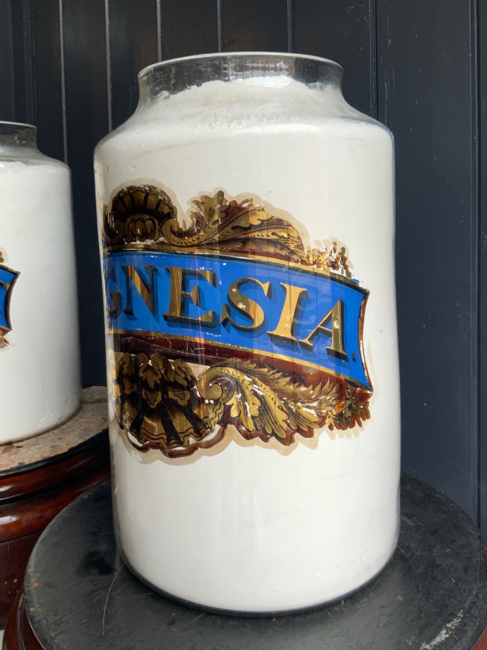A pair of cylindrical glass apothecary jars with ornate gold leaf and blue labels set on turned circular mahogany display bases. View V