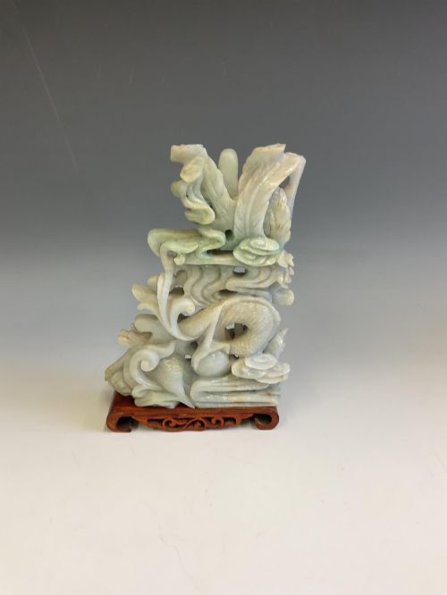 fig 1. A pale green carved jade sculpture depicting a dragon, phoenix, and double happiness character mounted on a dark carved hardwood stand.