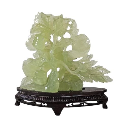 fig 1. A carved pale green Xiuyan jade sculpture depicting a lotus flower, foliage, and a central cash coin, displayed on a dark carved wooden stand.