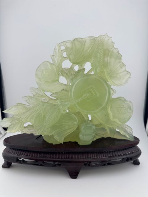 fig 2. A carved pale green Xiuyan jade sculpture depicting a lotus flower, foliage, and a central cash coin, displayed on a dark carved wooden stand.