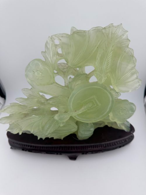 fig 3. A carved pale green Xiuyan jade sculpture depicting a lotus flower, foliage, and a central cash coin, displayed on a dark carved wooden stand.