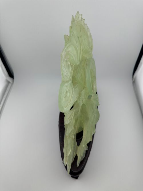 fig 4. A carved pale green Xiuyan jade sculpture depicting a lotus flower, foliage, and a central cash coin, displayed on a dark carved wooden stand.