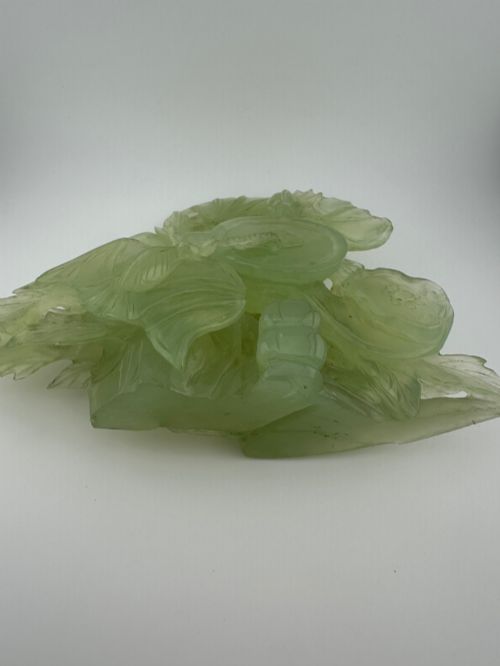 fig 9. A carved pale green Xiuyan jade sculpture depicting a lotus flower, foliage, and a central cash coin, displayed on a dark carved wooden stand.