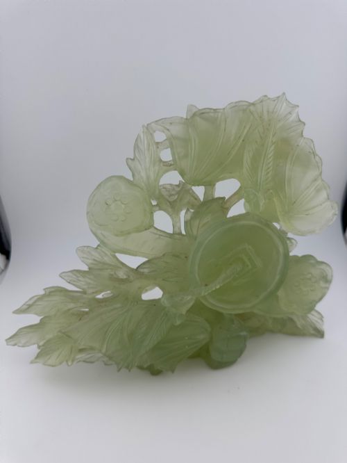 fig 10. A carved pale green Xiuyan jade sculpture depicting a lotus flower, foliage, and a central cash coin, displayed on a dark carved wooden stand.