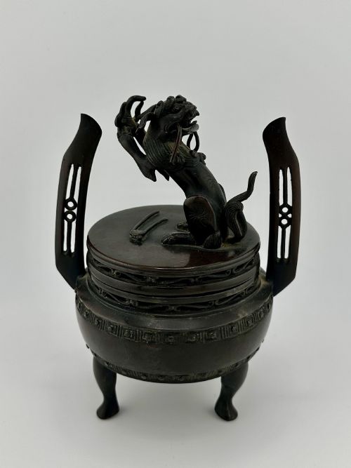 fig 1. A dark patinated bronze tripod censer featuring raised loop handles, a decorative meander border, and a fitted lid with a standing Qilin finial.