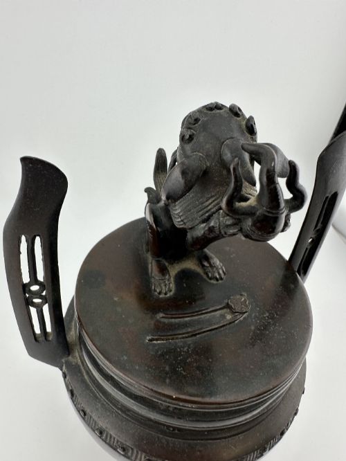 fig 2. A dark patinated bronze tripod censer featuring raised loop handles, a decorative meander border, and a fitted lid with a standing Qilin finial.
