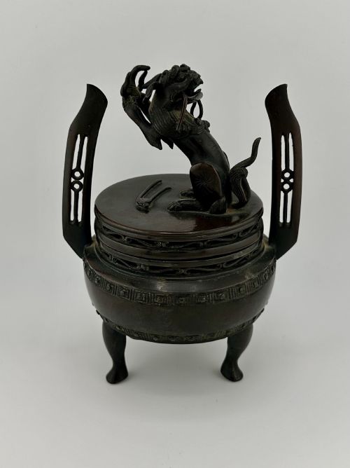 fig 4. A dark patinated bronze tripod censer featuring raised loop handles, a decorative meander border, and a fitted lid with a standing Qilin finial.