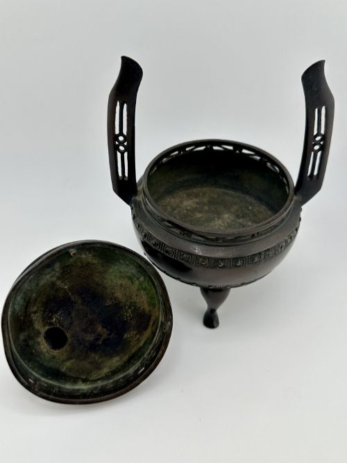 fig 8. A dark patinated bronze tripod censer featuring raised loop handles, a decorative meander border, and a fitted lid with a standing Qilin finial.