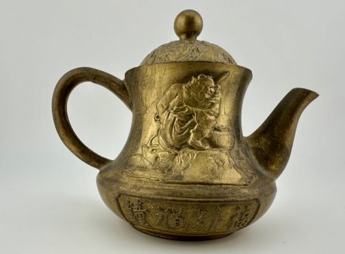 fig 1. A cast brass Chinese teapot featuring raised relief figures, traditional Chinese characters along the base, a curved spout, handle, and a round lid knob.