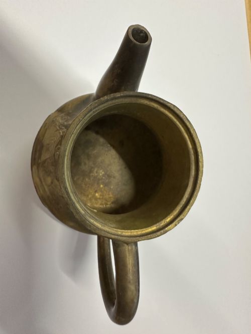 fig 2. A cast brass Chinese teapot featuring raised relief figures, traditional Chinese characters along the base, a curved spout, handle, and a round lid knob.