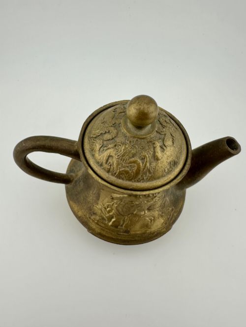 fig 3. A cast brass Chinese teapot featuring raised relief figures, traditional Chinese characters along the base, a curved spout, handle, and a round lid knob.