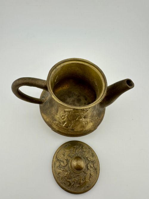 fig 4. A cast brass Chinese teapot featuring raised relief figures, traditional Chinese characters along the base, a curved spout, handle, and a round lid knob.