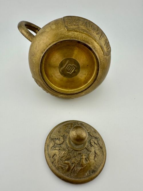 fig 5. A cast brass Chinese teapot featuring raised relief figures, traditional Chinese characters along the base, a curved spout, handle, and a round lid knob.