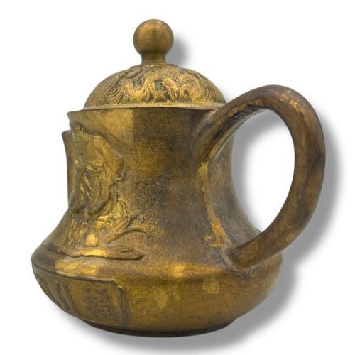 fig 6. A cast brass Chinese teapot featuring raised relief figures, traditional Chinese characters along the base, a curved spout, handle, and a round lid knob.