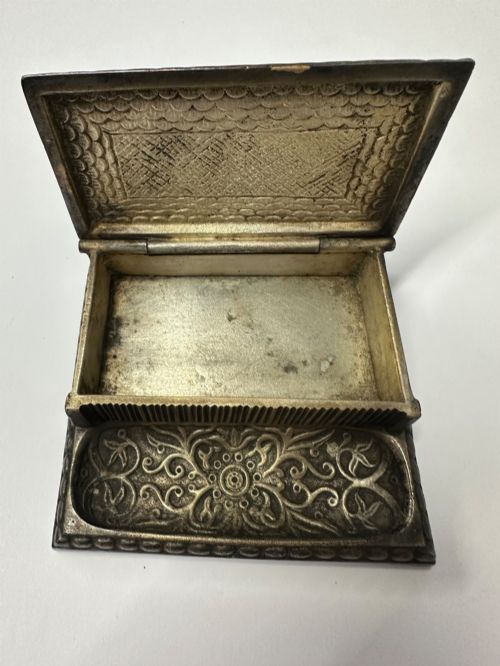 fig 1. An antique silvered brass desktop vesta box with a hinged floral embossed lid, ribbed striker strip, and front tray resting on a beaded base.