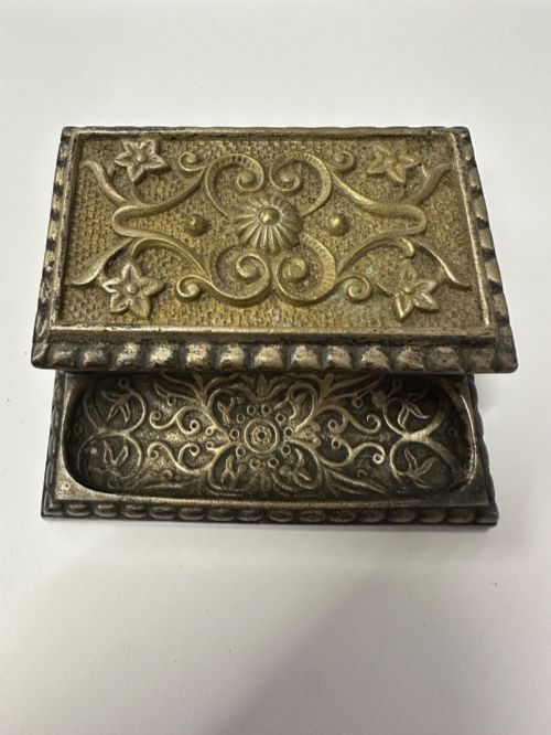 fig 3. An antique silvered brass desktop vesta box with a hinged floral embossed lid, ribbed striker strip, and front tray resting on a beaded base.