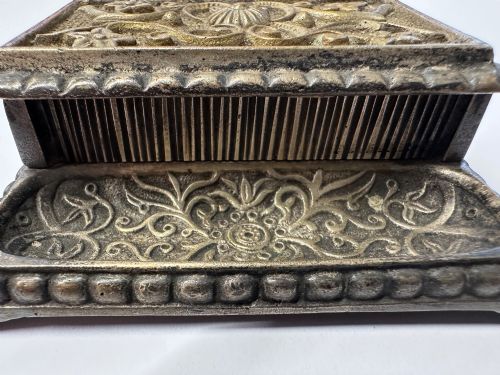 fig 5. An antique silvered brass desktop vesta box with a hinged floral embossed lid, ribbed striker strip, and front tray resting on a beaded base.