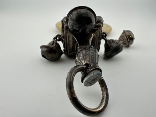 fig 1. An antique silver baby rattle featuring embossed metalwork, small attached bells, and a cream-coloured circular teether ring.
