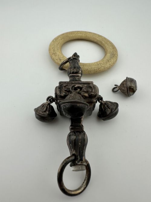 fig 2. An antique silver baby rattle featuring embossed metalwork, small attached bells, and a cream-coloured circular teether ring.