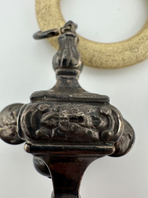 fig 4. An antique silver baby rattle featuring embossed metalwork, small attached bells, and a cream-coloured circular teether ring.