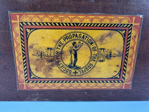 fig 2. A rectangular dark wooden church collection box featuring a coin slot on top and a printed yellow and red Society for the Propagation of the Gospel label on the front.