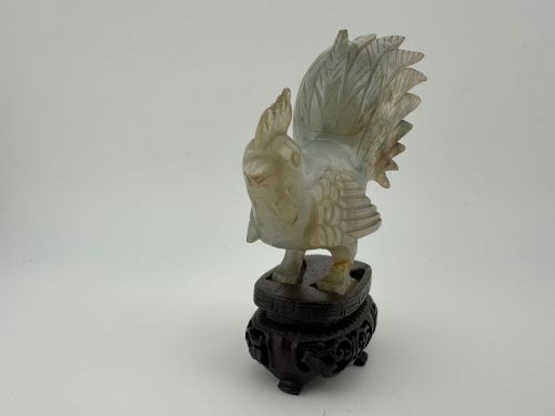 fig 1. A hand-carved pale green jade sculpture of a cockerel with fan-shaped tail feathers, displayed on a dark carved hardwood base.