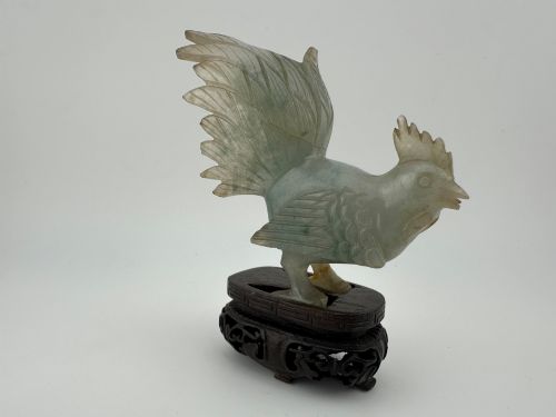 fig 2. A hand-carved pale green jade sculpture of a cockerel with fan-shaped tail feathers, displayed on a dark carved hardwood base.