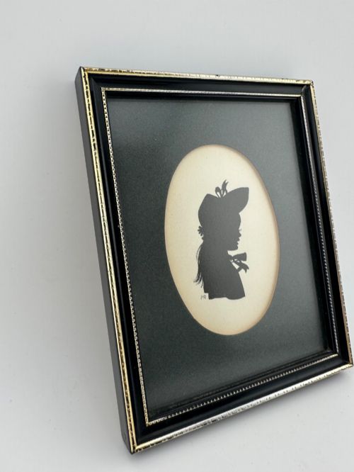 fig 2. A framed Art Deco silhouette portrait depicting a lady in profile with a decorative hat inside an oval cream mount surrounded by a black and gold frame.