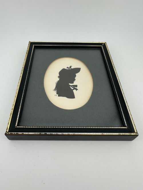 fig 3. A framed Art Deco silhouette portrait depicting a lady in profile with a decorative hat inside an oval cream mount surrounded by a black and gold frame.